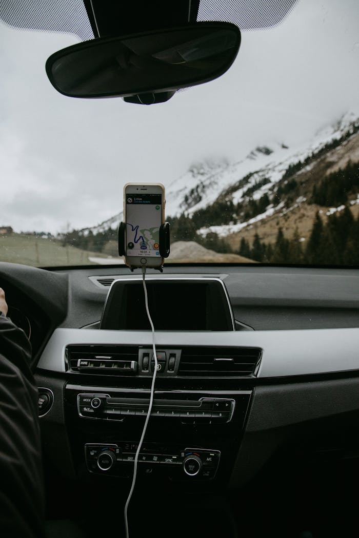 Driver in car with smartphone showing route due to GPS against snowy hill slope with coniferous trees and road